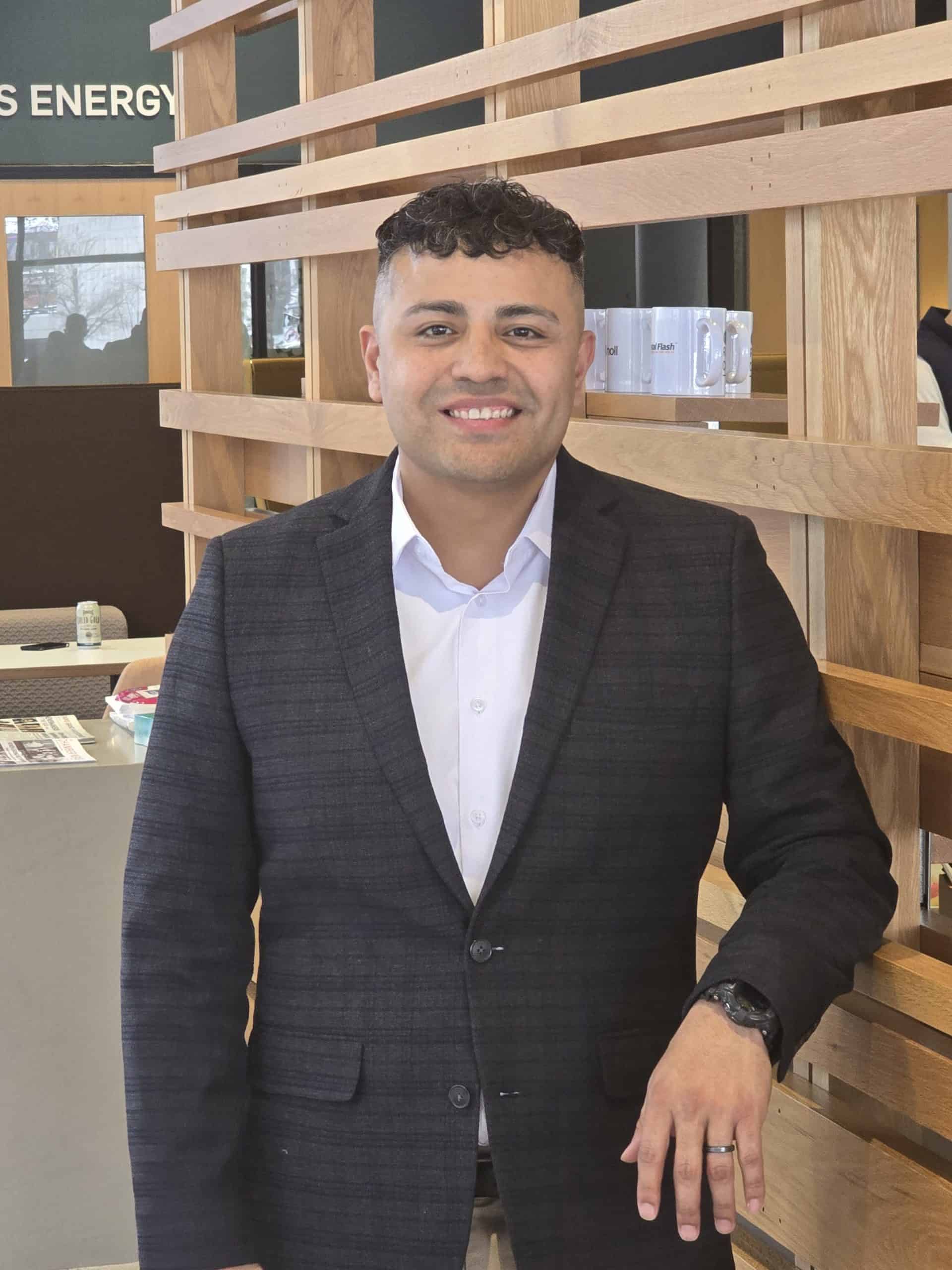 Johnny, a professional sales representative, standing in an office setting wearing a dark checked blazer and white shirt, smiling confidently.